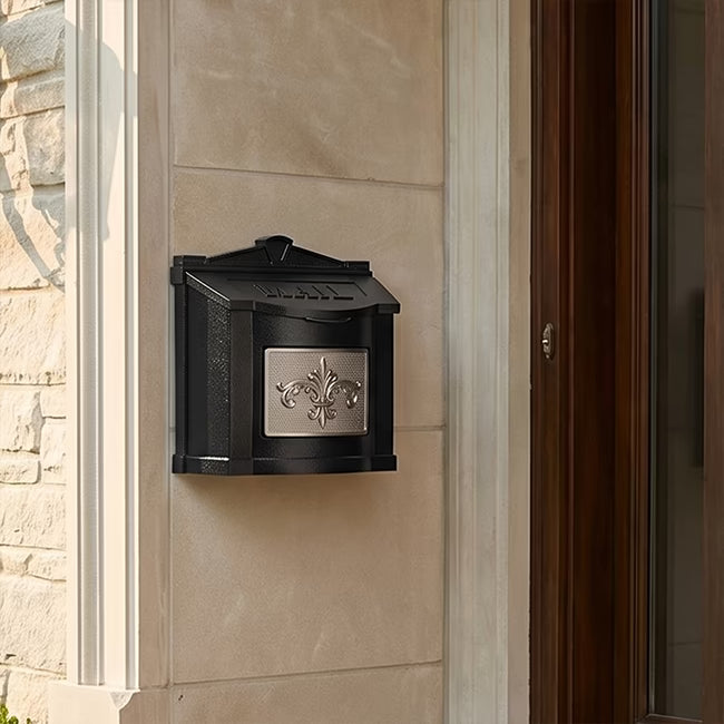 A black metal mailbox mounted on a beige stucco exterior wall next to a dark wood door, featuring a silver decorative metal front plate.