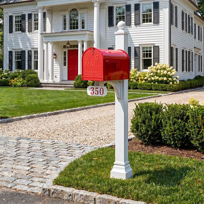 A classic white colonial-style house with black shutters and a red front door, set on a landscaped lawn with a red mailbox in the foreground under a blue sky.