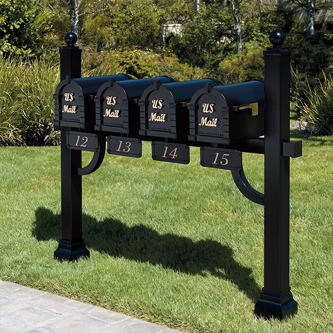 A black four-bell mailbox stand mounted on a grassy lawn, with gold lettering reading 'US Mail' on each mailbox and numbers 12 through 15 on the base plates. Trees and shrubs are visible in the background.